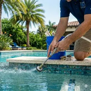 Person kneeling at the edge of a sunny backyard pool, using a pool skimmer to skim the water.