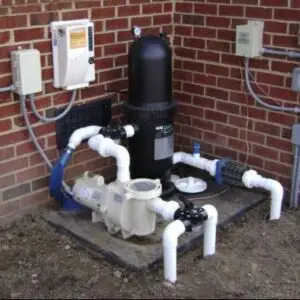 Outdoor pool filtration system on a concrete pad: a large black vertical filter with white PVC pipes and valves against a brick wall, plus control boxes nearby.