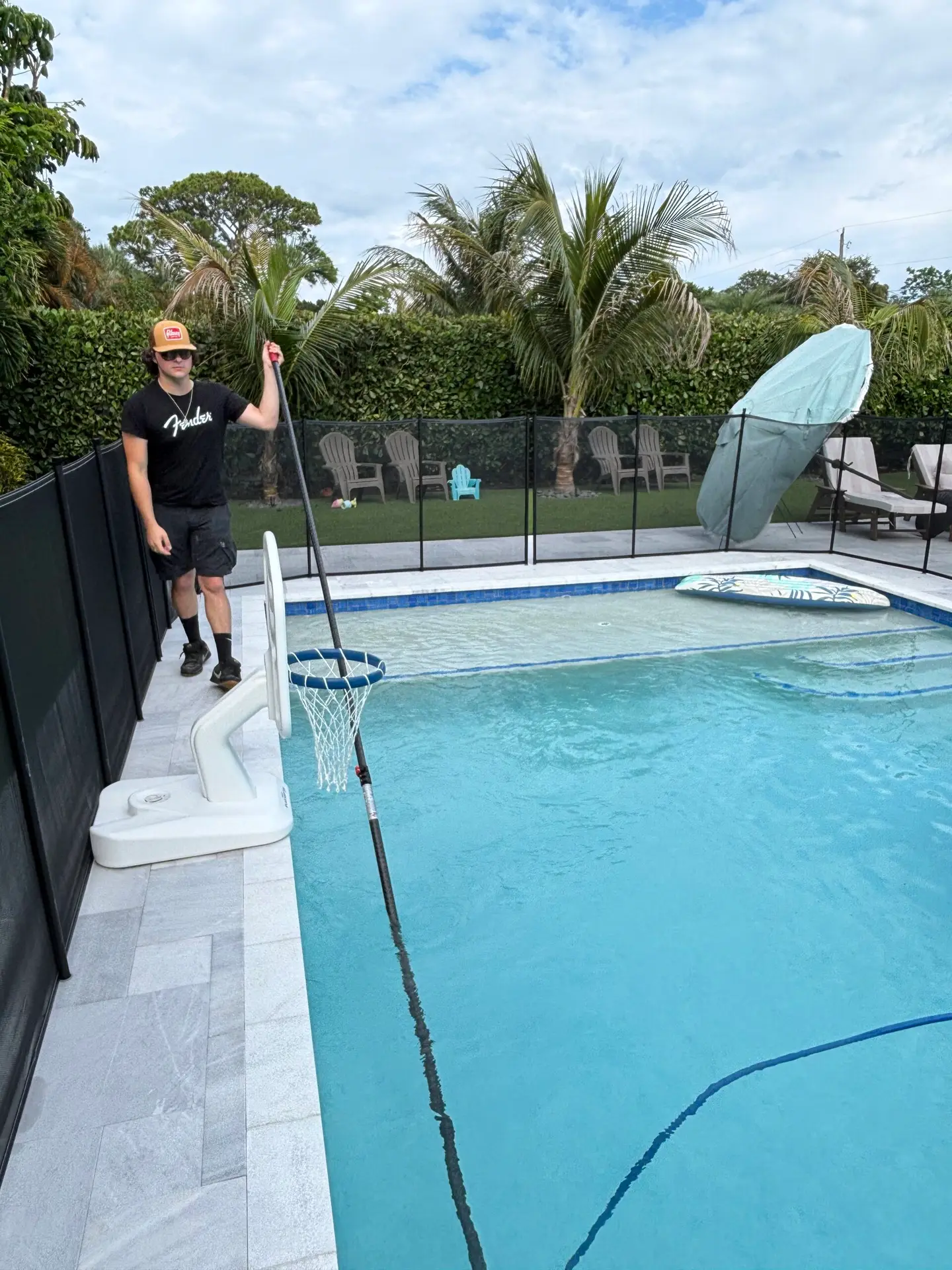 Man in a black t‑shirt and orange cap stands by a backyard pool, holding a long pole with a net over the water.